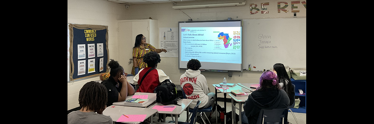 Standing women gives powerpoint presentation to seated students.
